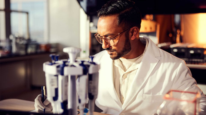 Male scientist, front side view, writing information on lab bench, pipettes in foreground